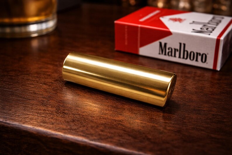 K&ouml;nnte beinhalten: Ein zylindrisches, goldfarbenes Metallobjekt liegt auf einer dunklen Holzoberfl&auml;che. Eine Packung Marlboro-Zigaretten mit rotem und wei&szlig;em Design befindet sich im Hintergrund. Die Szene ist mit warmem Licht beleuchtet.
