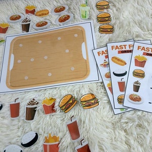 Puede incluir: Un juego de aprendizaje de comida r&aacute;pida con una bandeja de madera y varios art&iacute;culos de comida como hamburguesas, papas fritas, refrescos y hot dogs. El juego incluye tarjetas con el texto "Pedidos de comida r&aacute;pida" y n&uacute;meros que indican la cantidad de cada art&iacute;culo de comida.