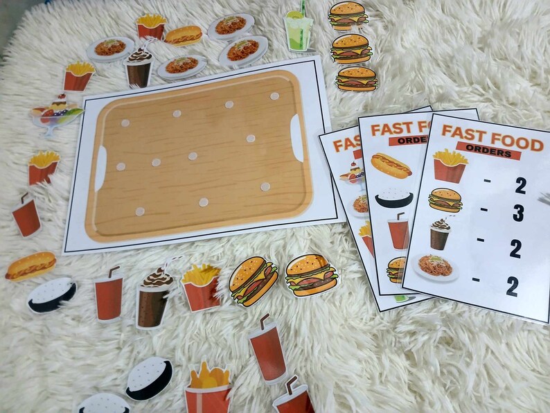 Puede incluir: Un juego de aprendizaje de comida r&aacute;pida con una bandeja de madera y varios art&iacute;culos de comida como hamburguesas, papas fritas, refrescos y hot dogs. El juego incluye tarjetas con el texto "Pedidos de comida r&aacute;pida" y n&uacute;meros que indican la cantidad de cada art&iacute;culo de comida.