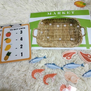 Puede incluir: Un conjunto colorido de tarjetas educativas con im&aacute;genes de frutas, verduras y otros alimentos. Las tarjetas tienen n&uacute;meros y las palabras "To Buy Lists" impresas en ellas. Tambi&eacute;n hay un cartel de mercado verde y blanco con una cesta de mimbre.
