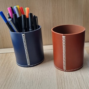 May include: Two leather pen holders, one blue and one brown, with white stitching. Both holders are cylindrical and have a diameter of approximately 7.5 cm.
