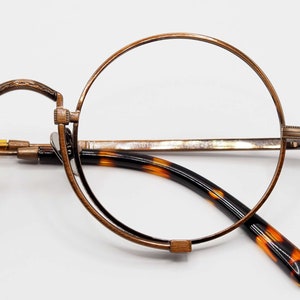 Puede incluir: Gafas redondas con montura de metal color bronce y lentes transparentes. Las patillas tienen un patrón de carey. Las gafas están diseñadas con una estética vintage.