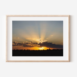 Op de afbeelding: Een foto van een zonsondergang boven een veld met een heldere oranje en gele lucht en wolken. De zon schijnt door de wolken en creëert een lichtstraal.