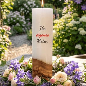 Könnte beinhalten: Eine hohe weiße Stumpenkerze mit brennender Flamme, einem Sockel aus Naturholz und dem schwarz-roten Text "Ihr eigenes Motiv." Die Kerze ist von einem Blumenarrangement aus violetten, rosafarbenen und cremefarbenen Blüten sowie grünen Blättern umgeben.