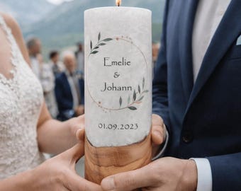May include: A cylindrical white wax candle with a lit flame, featuring a printed botanical wreath design, the names Emelie & Johann, and the date 01.09.2023. The base of the candle is made of natural light brown wood with a visible grain pattern.