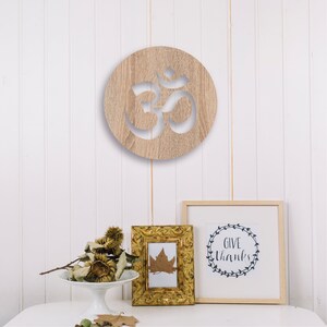 May include: A wooden wall decor with a cutout of the Om symbol. The decor is round and has a light brown wood grain. It is hanging on a white wall with a white wainscoting pattern. There are two framed pictures below the decor, one with a leaf and the other with the text "Give Thanks" in a wreath.
