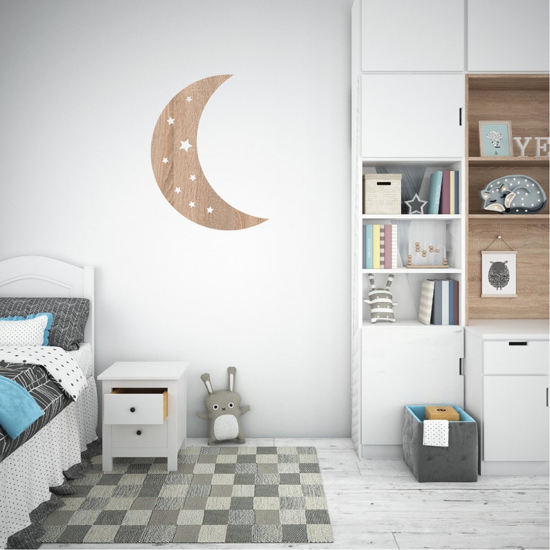 May include: A white wooden nightstand with two drawers, a white wooden bed frame with a white and black patterned duvet, a gray and white checkered rug, a white bookcase with shelves filled with books and toys, a gray storage bin, and a wooden crescent moon wall decor with white stars.