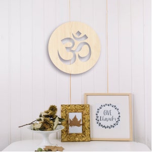 May include: A wooden wall decor featuring a circular cutout of the Om symbol. The symbol is carved into a light brown wooden circle. The circle is hanging on a white wall. There is a small framed print with the text "Give Thanks" and a small framed print with a leaf inside.