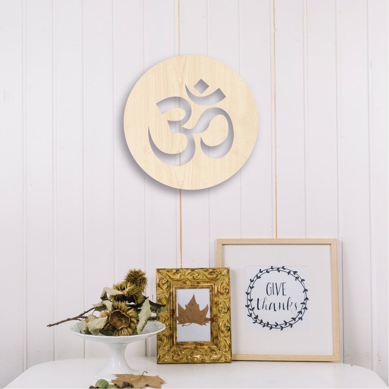 May include: A wooden wall decor featuring a circular cutout of the Om symbol. The symbol is carved into a light brown wooden circle. The circle is hanging on a white wall. There is a small framed print with the text "Give Thanks" and a small framed print with a leaf inside.