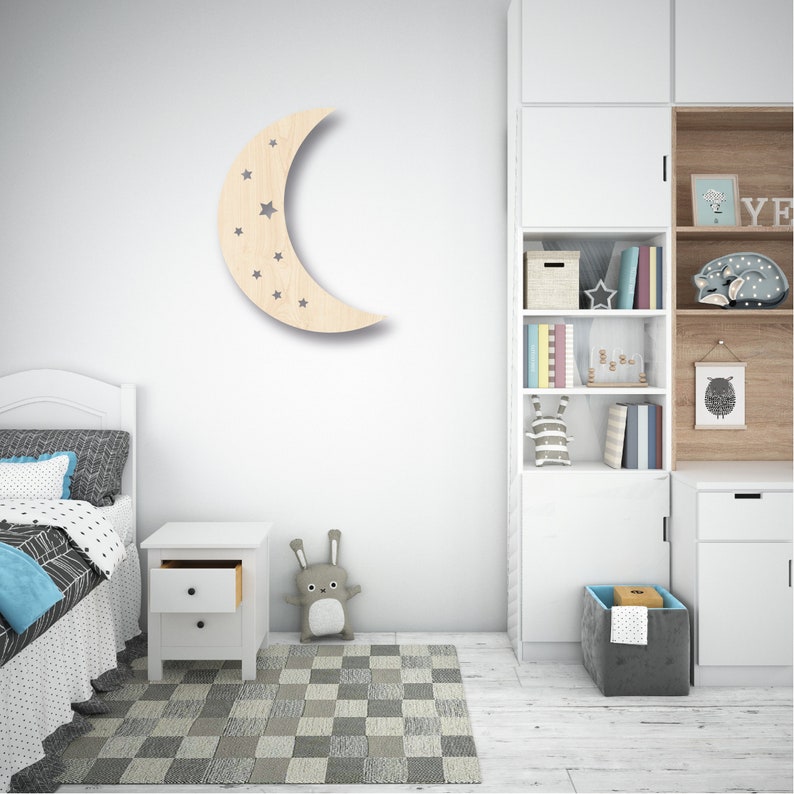 May include: A wooden crescent moon wall decor with cut-out stars. The moon is light brown and the stars are gray. The decor is hanging on a white wall in a child's bedroom.