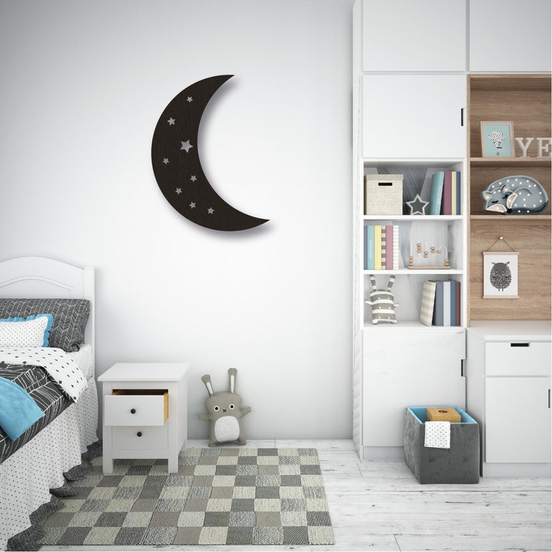 May include: A black wooden crescent moon wall decor with white stars. The moon is hanging on a white wall in a bedroom with a white nightstand, a white bed with a blue and white patterned bedspread, and a white bookcase with shelves filled with books and toys.