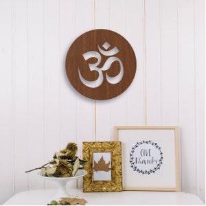 May include: A brown wooden circle with a white Om symbol cut out. The circle is hanging on a white wall. There are two framed pictures below the circle, one with a leaf and the other with the text "Give Thanks" in a wreath.