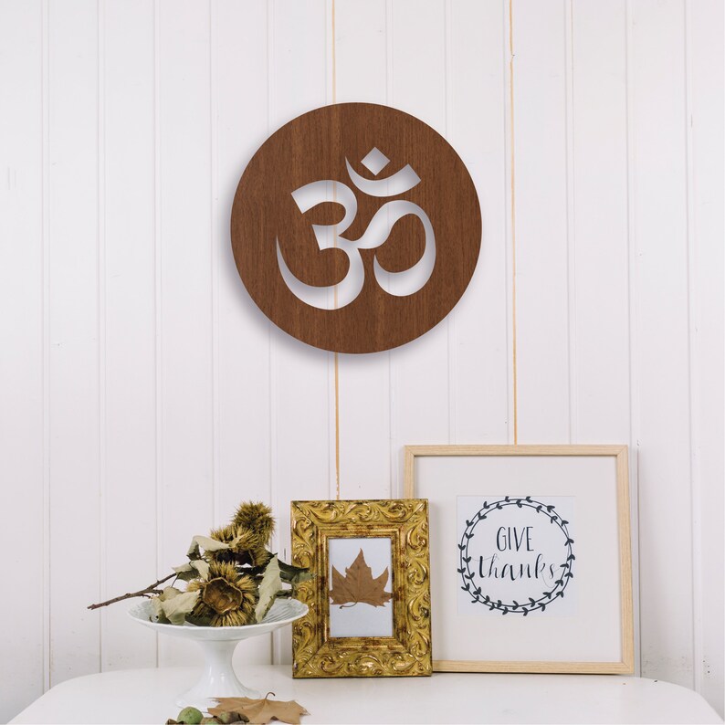 May include: A brown wooden circle with a white Om symbol cut out. The circle is hanging on a white wall. There are two framed pictures below the circle, one with a leaf and the other with the text "Give Thanks" in a wreath.