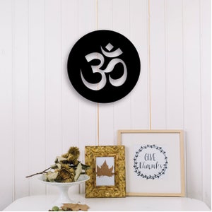 May include: Black and white circular wall decor with the Om symbol cut out. The decor is hanging on a white wall with a gold picture frame and a white picture frame with the text "Give Thanks" in a wreath design.