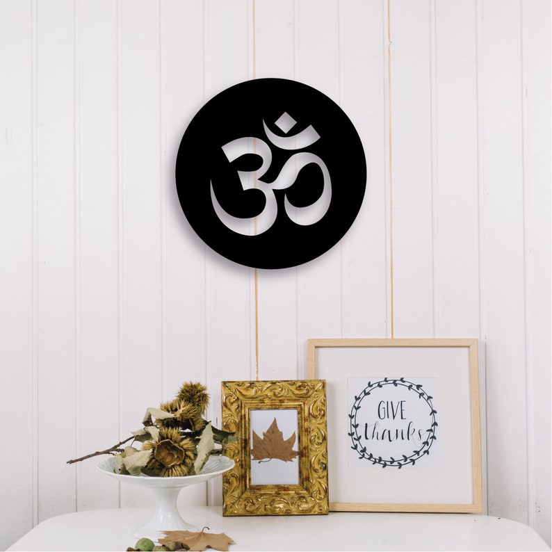 May include: Black and white circular wall decor with the Om symbol cut out. The decor is hanging on a white wall with a gold picture frame and a white picture frame with the text "Give Thanks" in a wreath design.