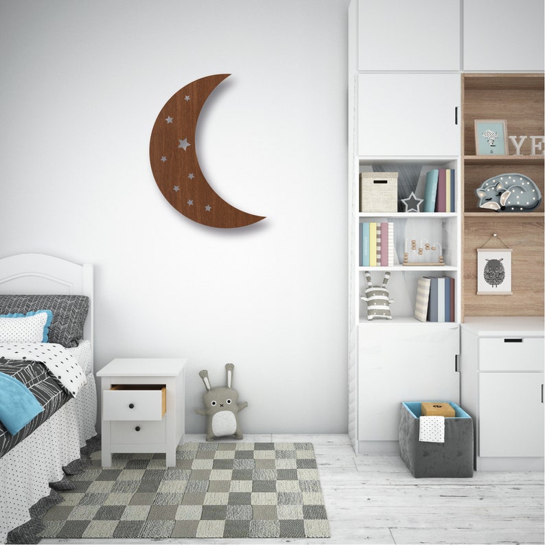 May include: A wooden crescent moon wall decor with a star design. The moon is brown and the stars are white. The decor is hanging on a white wall in a child's bedroom.