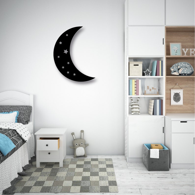 May include: Black crescent moon wall decor with white stars, hanging on a white wall in a child's bedroom. The room features a white bed with a black and white patterned duvet, a white nightstand, and a white bookshelf with books and toys.