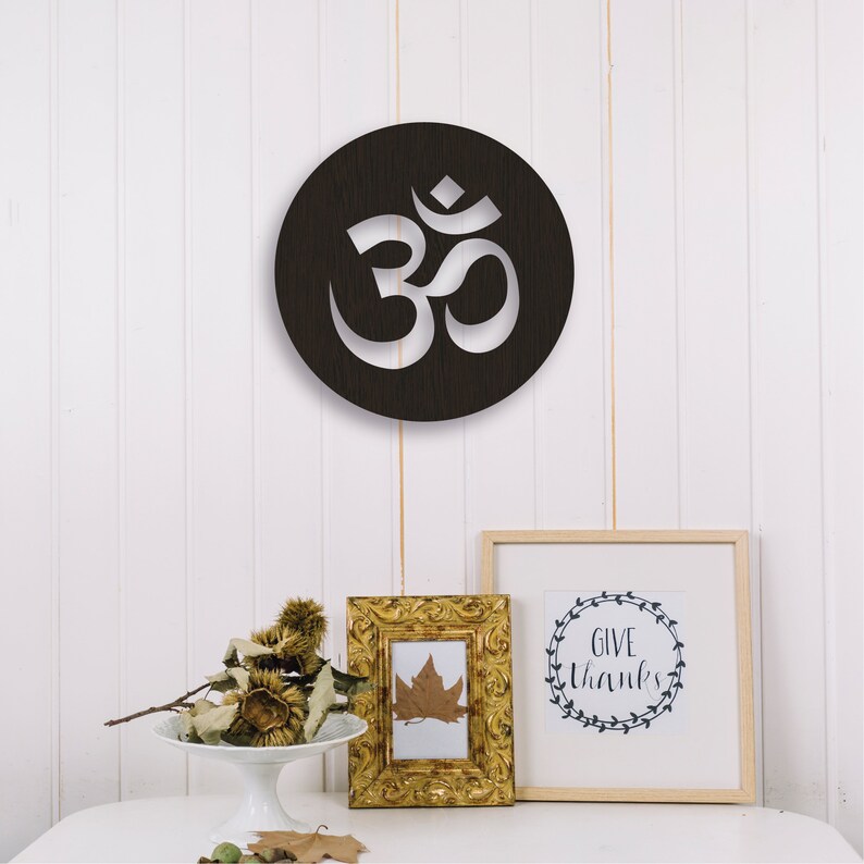 May include: A black circular wooden wall decor with a white cutout of the Om symbol. The decor is hanging on a white wall with two framed pictures below it.