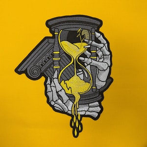 May include: An embroidered patch featuring a skeletal hand holding a dark gray hourglass with yellow sand leaking from the bottom. A classical architectural column fragment is positioned behind the hourglass, all outlined in black thread.