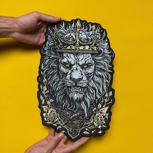 Pode incluir: Patch preto com uma ilustração detalhada de um leão usando uma coroa de ouro. O leão tem uma expressão feroz com dentes visíveis e é cercado por elementos florais. O patch é segurado contra um fundo amarelo.