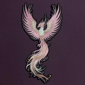 Könnte beinhalten: Gestickter Aufnäher mit einem Phönix mit ausgebreiteten Flügeln. Die Federn sind rosa, gold und weiß, mit schwarzer Umrandung. Der Aufnäher befindet sich auf einem dunkelvioletten Hintergrund, der das detaillierte Design zeigt.
