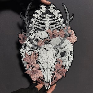 Peut inclure: Écusson brodé représentant un crâne de cerf avec des bois, des crânes, une cage thoracique et des fleurs roses. Le patch a une bordure noire et est tenu par les mains d'une personne. Le motif est détaillé avec du fil blanc, gris et rose.