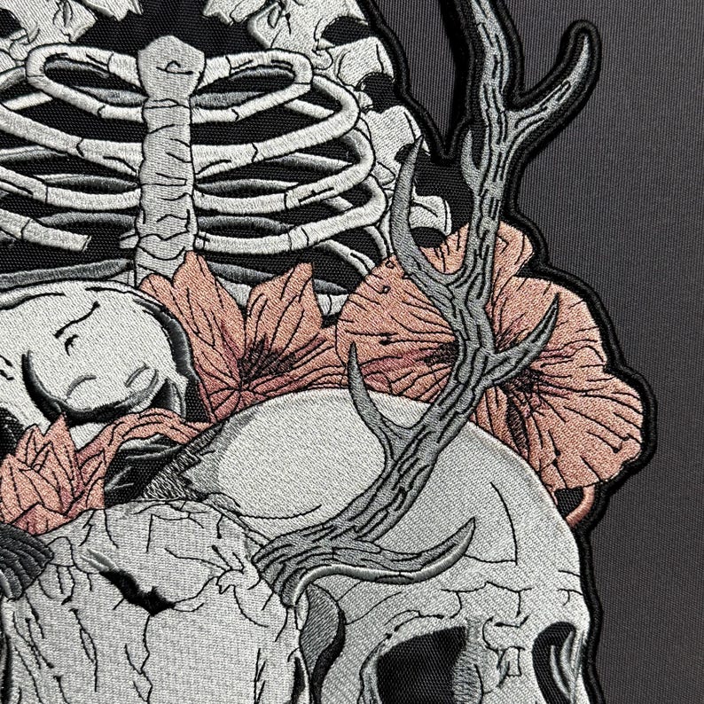 May include: Embroidered patch featuring a skull with antlers, surrounded by flowers and a rib cage. The design incorporates shades of gray, peach, and black, creating a gothic or alternative aesthetic. The patch is likely for clothing or accessories.
