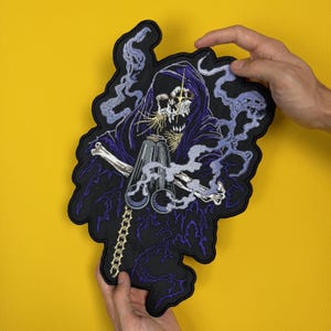 May include: Embroidered patch featuring a skeletal figure in a dark cloak, holding a chain and a metallic object. The design incorporates purple and white smoke-like elements against a black background. The patch is held against a yellow background.