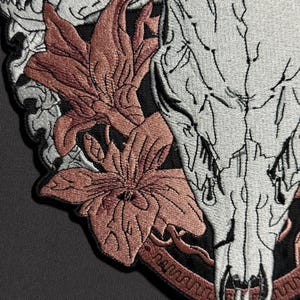May include: Embroidered patch featuring a detailed design of a skull, flowers, and decorative elements. The skull is primarily white with black outlines, while the flowers are a muted reddish-brown. The patch has a black border and is likely for clothing or accessories.
