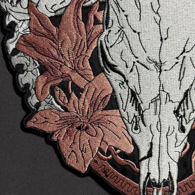 May include: Embroidered patch featuring a detailed design of a skull, flowers, and decorative elements. The skull is primarily white with black outlines, while the flowers are a muted reddish-brown. The patch has a black border and is likely for clothing or accessories.
