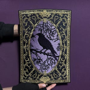 Könnte beinhalten: Ein rechteckiger, schwarz-violetter, gestickter Aufnäher mit einem Raben, der auf einem Ast in einem ovalen Rahmen sitzt. Der Rahmen und der umliegende Rand haben aufwendige goldene Details. Der Hintergrund ist einfarbig violett.