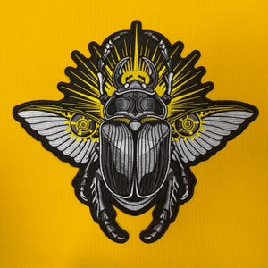 May include: An embroidered patch featuring a stylized beetle with metallic silver and black thread detailing. The insect has expansive wings with gear-like circular motifs and a radiant yellow sunburst pattern emanating from behind its head.
