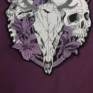 Op de afbeelding: Een geborduurde patch met een centrale witte herten-schedel met gewei, geflankeerd door twee menselijke schedels aan de zijkanten. Het ontwerp is geaccentueerd met paarse leliebloemen en een cirkelvormige rand op een donkerpaarse stoffen ondergrond.