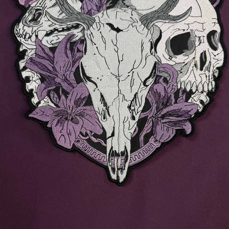Op de afbeelding: Een geborduurde patch met een centrale witte herten-schedel met gewei, geflankeerd door twee menselijke schedels aan de zijkanten. Het ontwerp is geaccentueerd met paarse leliebloemen en een cirkelvormige rand op een donkerpaarse stoffen ondergrond.