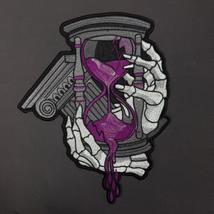 Puede incluir: Un parche bordado que muestra una mano esquelética sosteniendo un reloj de arena roto con arena morada goteando por la parte inferior. El diseño incluye un elemento de columna arquitectónica gris y está delineado con hilo negro sobre un fondo oscuro.