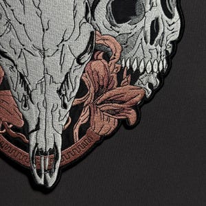 May include: Embroidered patch featuring a detailed design of a skull and a deer skull, with floral accents in shades of gray, black, and rust. The patch has a textured appearance, suitable for application on clothing or accessories.