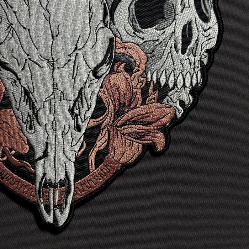May include: Embroidered patch featuring a detailed design of a skull and a deer skull, with floral accents in shades of gray, black, and rust. The patch has a textured appearance, suitable for application on clothing or accessories.
