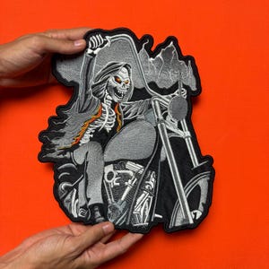 Op de afbeelding: Geborduurde patch met een skeletmotorrijder op een motorfiets. De patch is voornamelijk zwart en grijs, met oranje en gele accenten op de borst en cape van het skelet. De motorrijder houdt een zeis vast en het ontwerp heeft een gedetailleerde, gotische esthetiek.