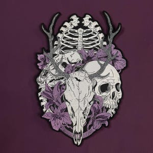 Op de afbeelding: Een geborduurde patch met een centrale herten schedel met gewei, geflankeerd door een menselijke schedel en een ribbenkast. Het ontwerp is geaccentueerd met paarse leliebloemen en zwarte omlijningen, wat een gedetailleerde compositie vormt.