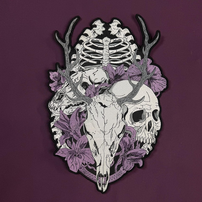 Op de afbeelding: Een geborduurde patch met een centrale herten schedel met gewei, geflankeerd door een menselijke schedel en een ribbenkast. Het ontwerp is geaccentueerd met paarse leliebloemen en zwarte omlijningen, wat een gedetailleerde compositie vormt.