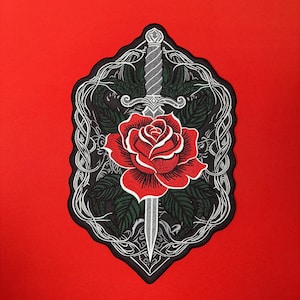 Puede incluir: Un parche bordado rectangular que presenta una rosa roja central atravesada por una daga plateada. El diseño está enmarcado por hojas de color verde oscuro y líneas blancas entrelazadas sobre un fondo negro, todo sobre una superficie roja.