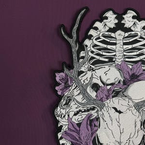 Op de afbeelding: Een geborduurde patch met een witte dierenschedel met gewei, een ribbenkast en paarse bloemen. Het ontwerp gebruikt zwart, wit en paars garen op een donkerpaarse stoffen achtergrond, wat een gedetailleerde grafische illustratie vormt.