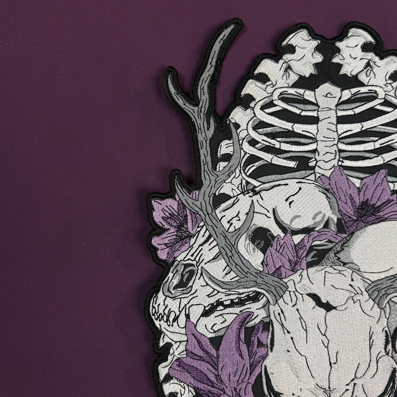 Op de afbeelding: Een geborduurde patch met een witte dierenschedel met gewei, een ribbenkast en paarse bloemen. Het ontwerp gebruikt zwart, wit en paars garen op een donkerpaarse stoffen achtergrond, wat een gedetailleerde grafische illustratie vormt.
