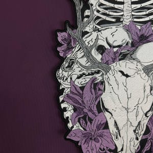 Op de afbeelding: Een geborduurde patch met een witte dierenschedel met gewei, omringd door paarse bloemen en een gedeeltelijke menselijke ribbenkast. Het ontwerp is uitgevoerd in zwart-wit garen op een zwarte achtergrond, met een donkerpaarse stoffen rand.