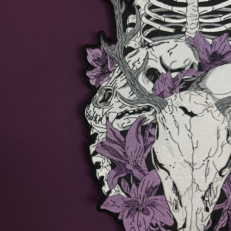 Op de afbeelding: Een geborduurde patch met een witte dierenschedel met gewei, omringd door paarse bloemen en een gedeeltelijke menselijke ribbenkast. Het ontwerp is uitgevoerd in zwart-wit garen op een zwarte achtergrond, met een donkerpaarse stoffen rand.