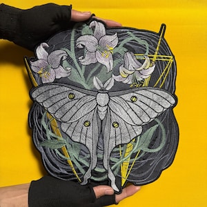 May include: Embroidered patch featuring a large grey moth with detailed wing patterns, set against a backdrop of stylised flowers and foliage. The design incorporates geometric shapes and yellow accents, all on a black background.