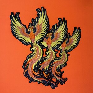 Op de afbeelding: Drie geborduurde phoenix-patches. Elke patch heeft een phoenix met gele, oranje en rode details op een zwarte achtergrond. De patches zijn geplaatst tegen een oranje achtergrond. De phoenixen hebben uitgespreide vleugels.