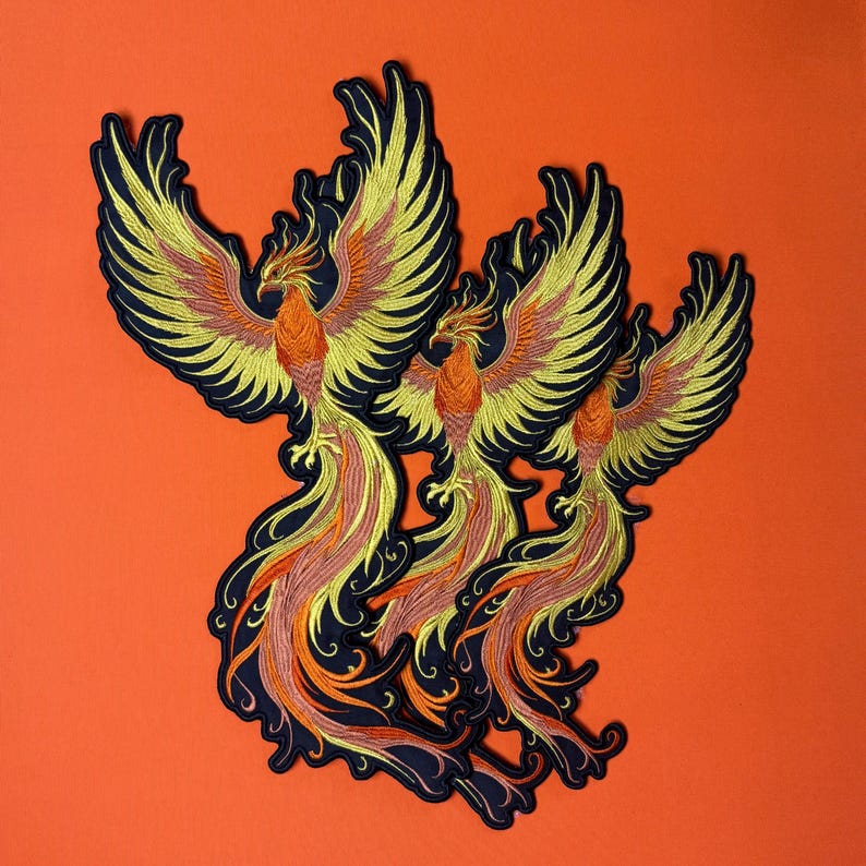 Op de afbeelding: Drie geborduurde phoenix-patches. Elke patch heeft een phoenix met gele, oranje en rode details op een zwarte achtergrond. De patches zijn geplaatst tegen een oranje achtergrond. De phoenixen hebben uitgespreide vleugels.