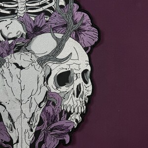 Op de afbeelding: Een geborduurde patch op donkerpaarse stof met een witte menselijke schedel en een dierenschedel met gewei, omringd door paarse bloemen en een gedeeltelijke ribbenkast. Het ontwerp gebruikt zwarte omlijningen om de details te defini&euml;ren.
