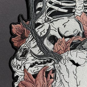 May include: Embroidered patch featuring a detailed skull with antlers and floral accents in shades of white, gray, and coral. The patch has a black border and is likely for jackets or bags.